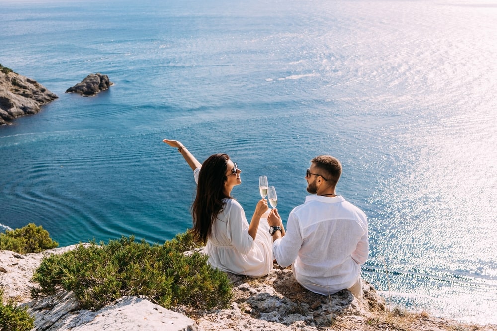 A couple in love celebrates their engagement on the seashore. A beautiful couple drinks champagne by the sea. Honeymoon trip. Lovers on the beach. Wedding travel. Couple on vacation.