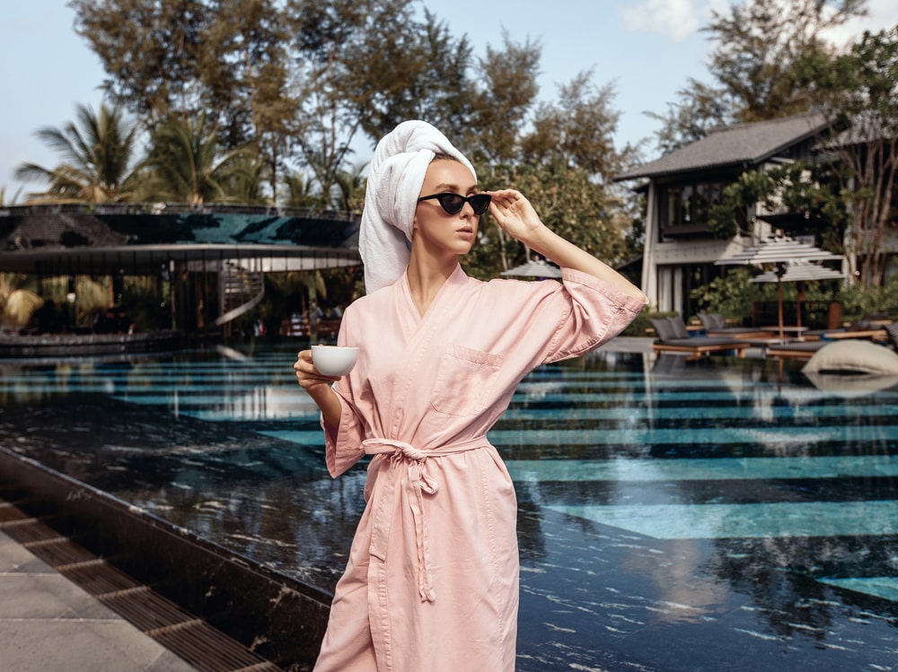 Portrait of a rich and confident young girl wearing a bathrobe, sunglasses and a towel on her head, drinking a cup of morning coffee and enjoying the morning in front of the pool and villa.