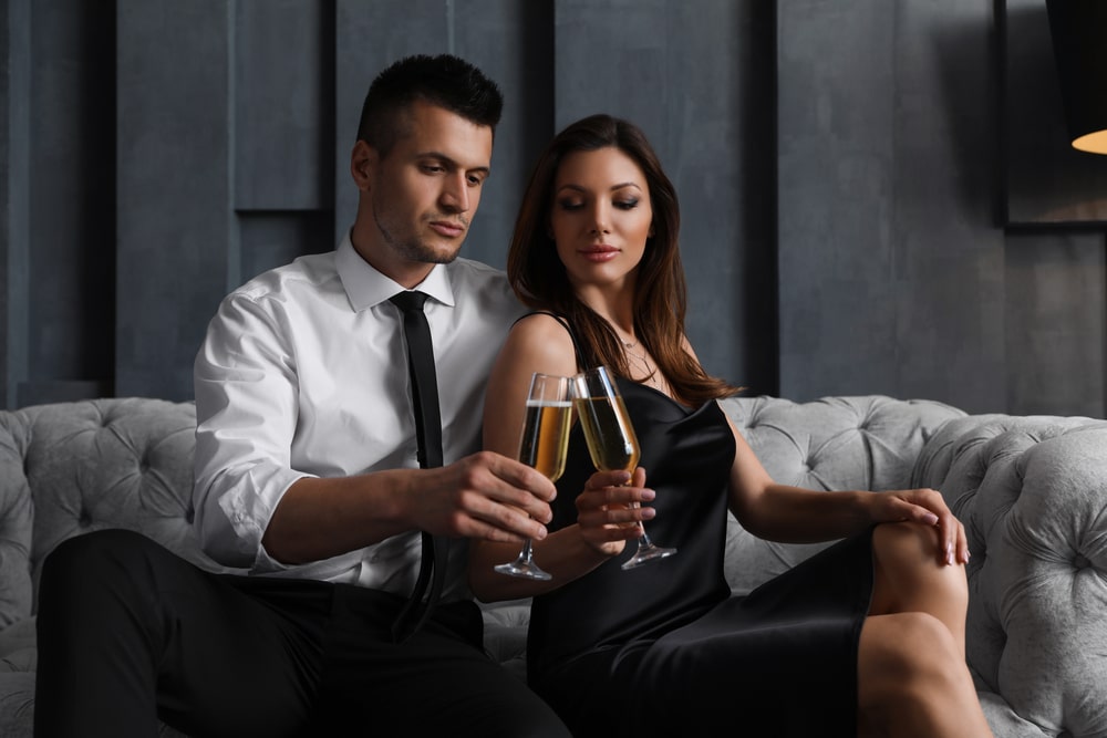 Beautiful couple who met on millionaire dating website with glasses of champagne on sofa indoors. Luxury lifestyle,