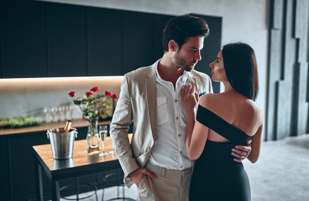 A chic couple posing in a contemporary kitchen. The dapper, bearded man wears a suit, while the stunning woman dons an elegant black dress, both radiating elegance in their home setting.