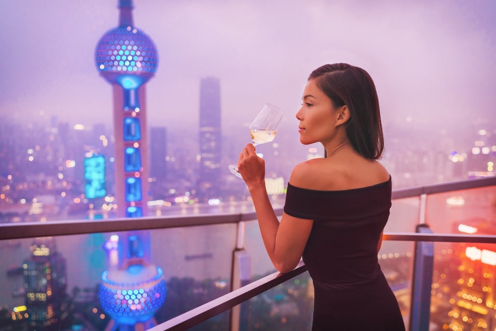 Femme tenant un verre de vin blanc dans une robe noire, debout près de la clôture sur la terrasse et réfléchissant à la façon de parler à un sugar daddy et de l'impressionner.