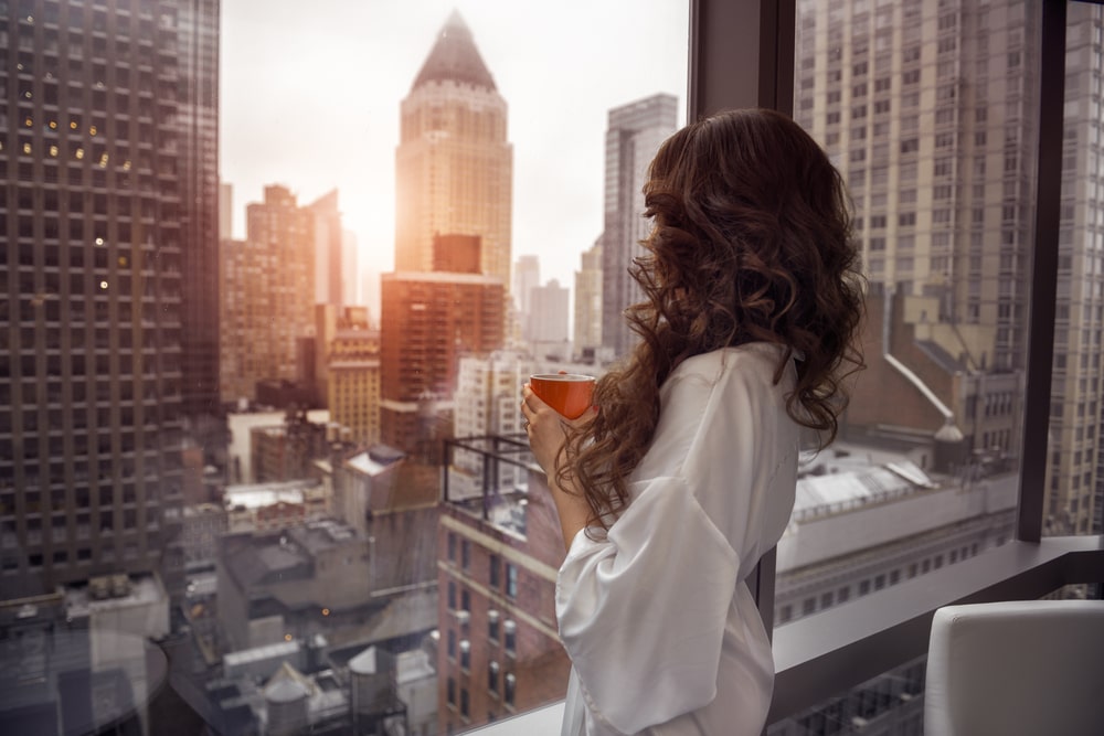 a woman stands by the window in a luxury neighborhood and looks at the metropolis, holding a salt shaker with a coffee in her hand