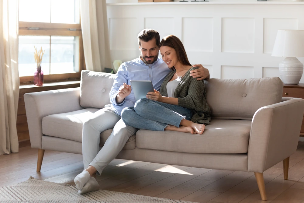 Glückliches junges Paar sitzt gemütlich auf dem Sofa, schaut gemeinsam auf ein Tablet und genießt entspannte Zeit zu Hause.