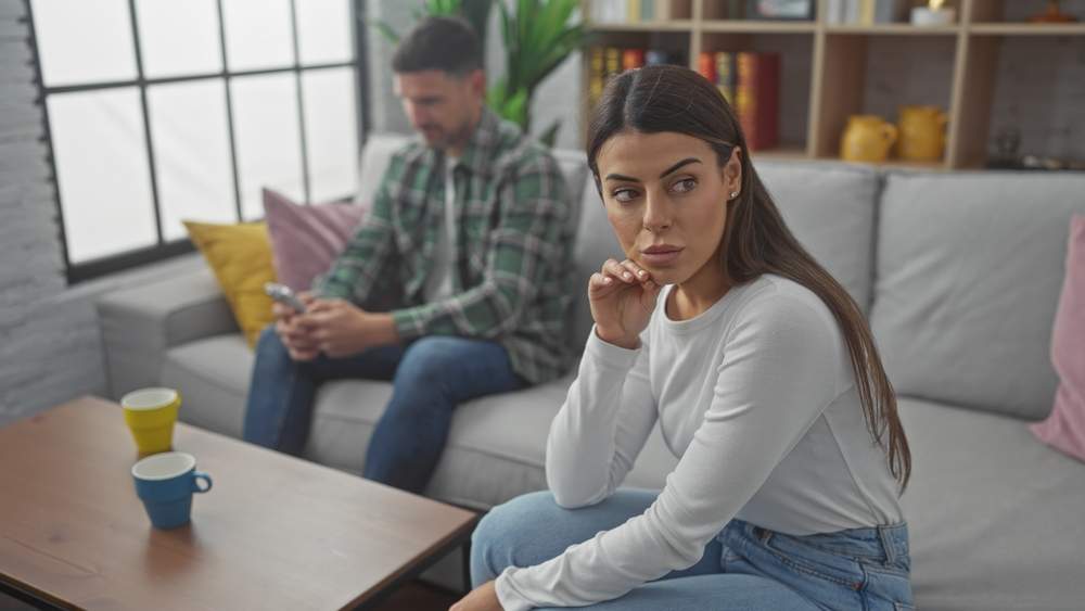 Frau sitzt nachdenklich auf dem Sofa waehrend ein Mann im Hintergrund aufs Handy schaut als Symbol fuer emotionale Distanz und Fremdverliebtsein.